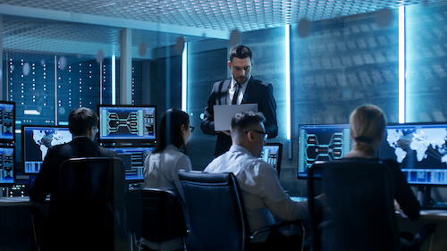 Professional IT Engineers Working in System Control Center Full of Monitors and Servers. Supervisor Holds Laptop and Holds a Briefing. Possibly Government Agency Conducts Investigation.