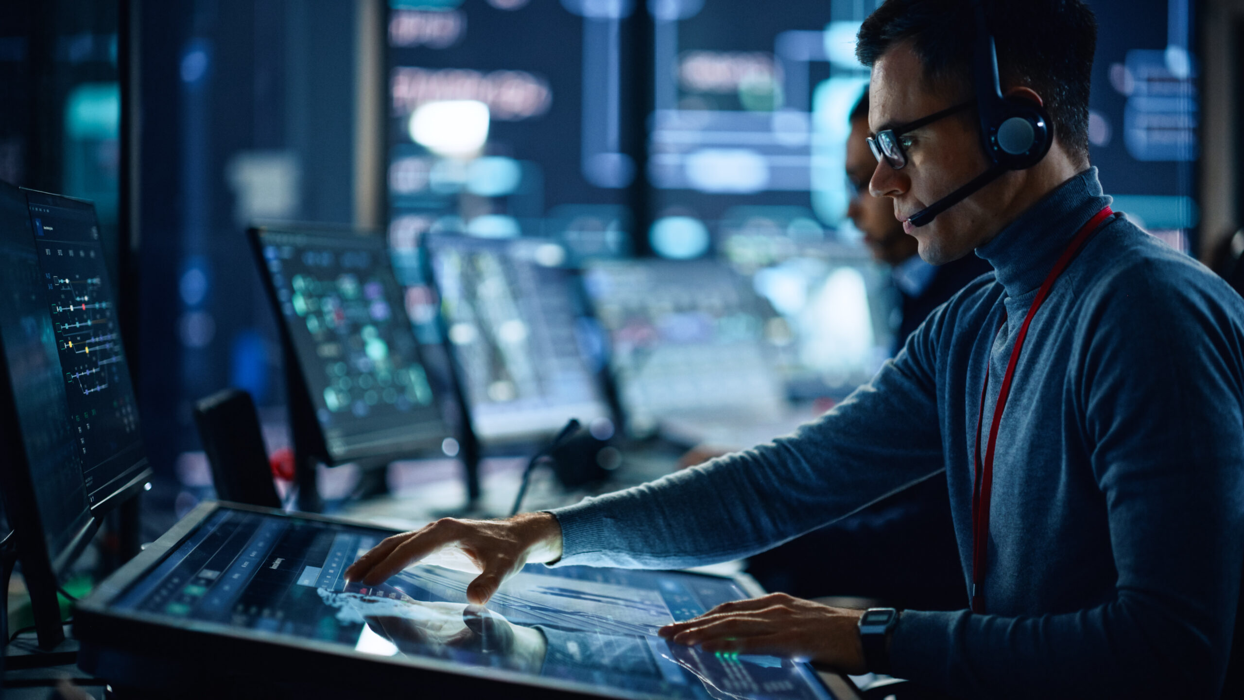 Portrait of Professional IT Technical Support Specialist Working on Computer in Monitoring Control Room with Digital Screens. Employee Wears Headphones with Mic and Talking on a Call.